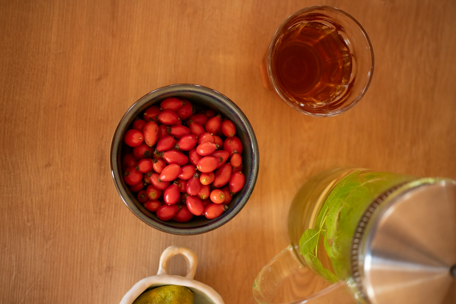 A glass of herbal tea beside a teapot and a bowl of red rose hips